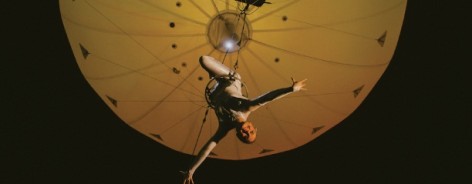 Photo of a trapeze artist hanging beneath a yellowy balloon