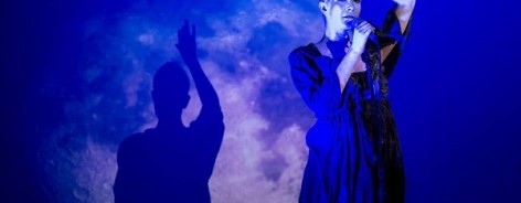Photo of a silver painted woman performing in front of the image of a moon