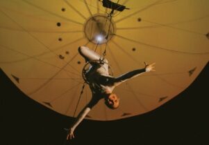 Photo of a trapeze artist hanging beneath a yellowy balloon