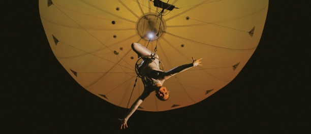 Photo of a trapeze artist hanging beneath a yellowy balloon