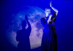 Photo of a silver painted woman performing in front of the image of a moon