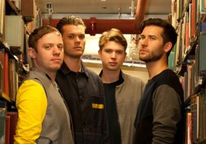 Photo of the band members of Everything Everything between the library's bookshelves.