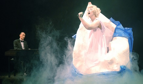 Photo of a woman in a pink dress singing with a man on piano behind