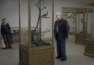 Still from the film with a man looking at a bird in a case