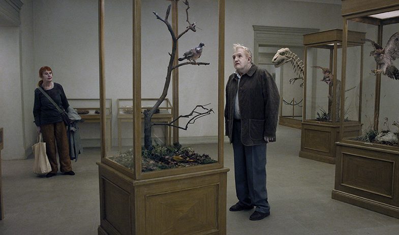 Still from the film with a man looking at a bird in a case