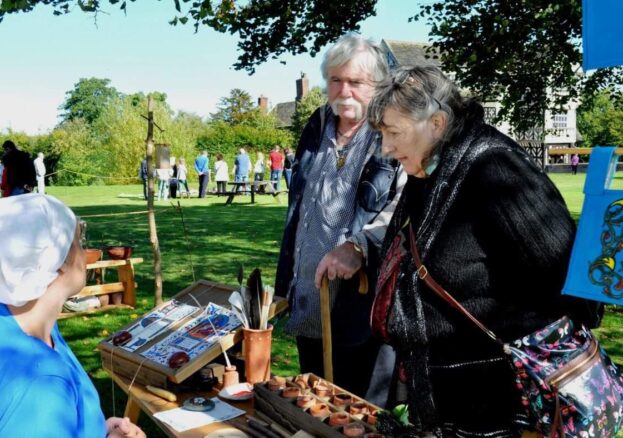 Living history event at Little Moreton Hall. A lady in period dress sits at a desk talking to visitors in the outdoors.