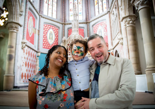 Happy Sundaze at Manchester Monastery. Young child with face paint is held by a man and woman on either side inside the monastery.
