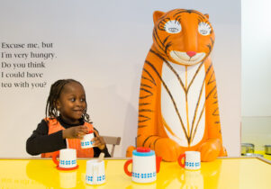 Half term fun - Young girl sat next to the tiger as part of the The Tiger who came to tea and the adventures of mog the forgetful cat exhibition