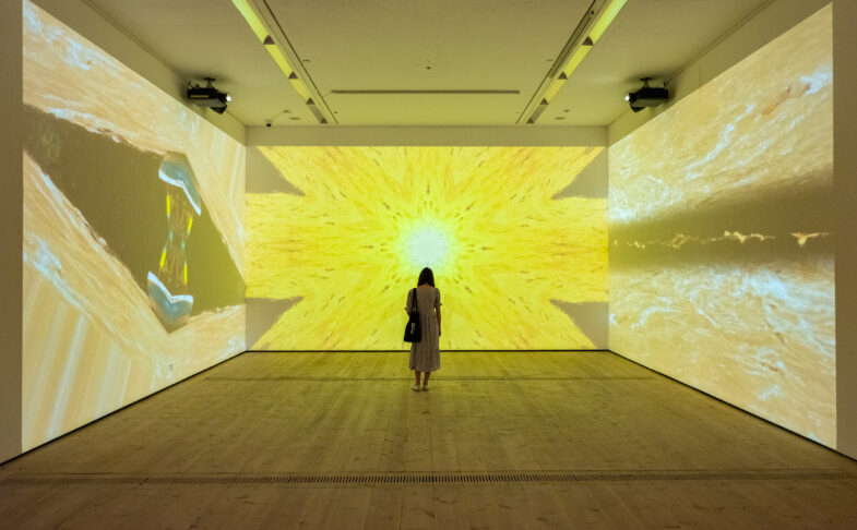 Patron Mono by Carolina Caycedo, image depicts a figure standing in a room with a three channel video emitting a yellow light