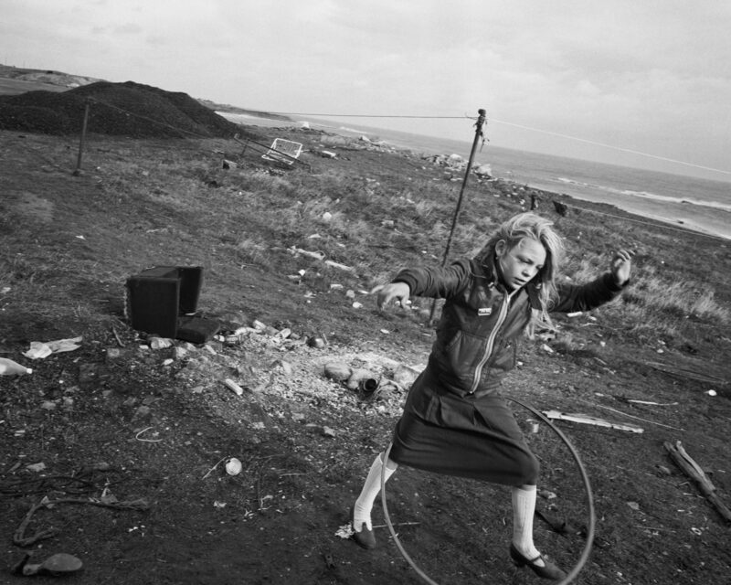 © Chris Killip Photography Trust/Magnum Photos, courtesy of Martin Parr Foundation. Black and white photo depicting young girl playing with a hula hoop in what looks like a wasteland by the sea