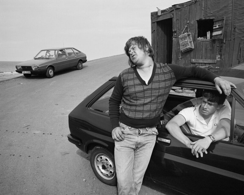 © Chris Killip Photography Trust/Magnum Photos, courtesy of Martin Parr Foundation. Black and white photo depicts two men, one leaning on a car, the other inside with the window open.