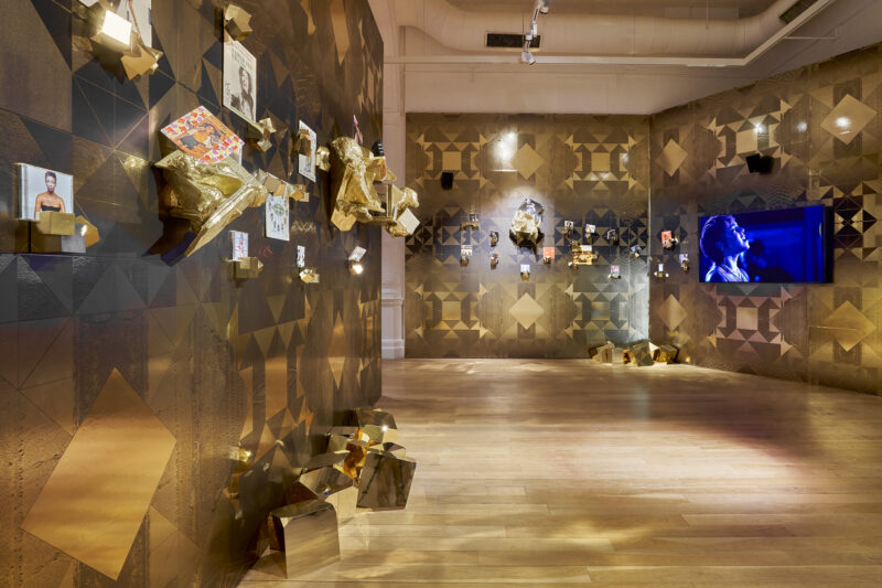 Exhibition view of Sonia Boyce: Feeling Her Way featuring gold, patterned wallpaper and a video screen