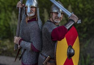 two men pose in chain mail. They are wearing metal helmets, one is holding an axe in the air and the other is holding a sowrd in the air. They are facing the camera.