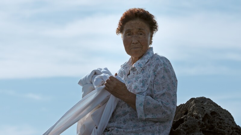 Still from 'Halmang', a video artwork by Jane Jin Kaisen. An elderly woman with short brown hair leans against a rock on Jeju Island, south of the Korean peninsula. Looking at the viewer with a neutral expression, she wears a long sleeved blouse with a floral pattern and is holding a long white cloth.