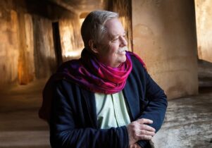 Image of Armistead Maupin looking to thier side, wearing a navy blue cardigan and magenta scarf
