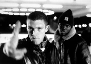 Two men look at the camera, one points his fingers as a gun at the viewer.