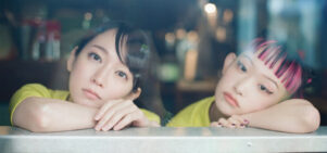 Two Japanese girls lean their heads on their hands looking out of a window, bored.