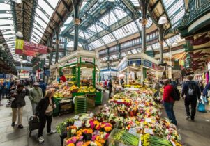 A photograph of the huge 1904 Hall in Kirkgate Market Leeds. The image shows a wide-angle shot from low-down capturing the colours of the florists and fruit and veg stall in the foreground.