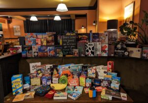 Lots of different boardgames arranged on a table and the shelf behind the table.