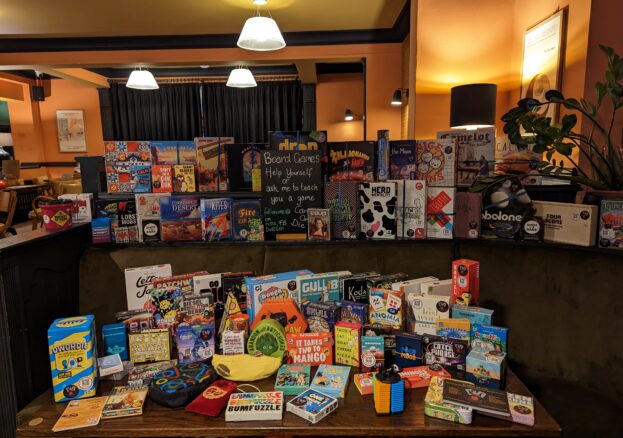 Lots of different boardgames arranged on a table and the shelf behind the table.
