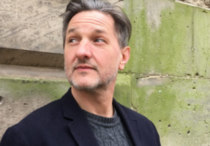 Image of author C. D. Rose looking to the side, wearing a black coat and grey jumper, stood in front of a wall.