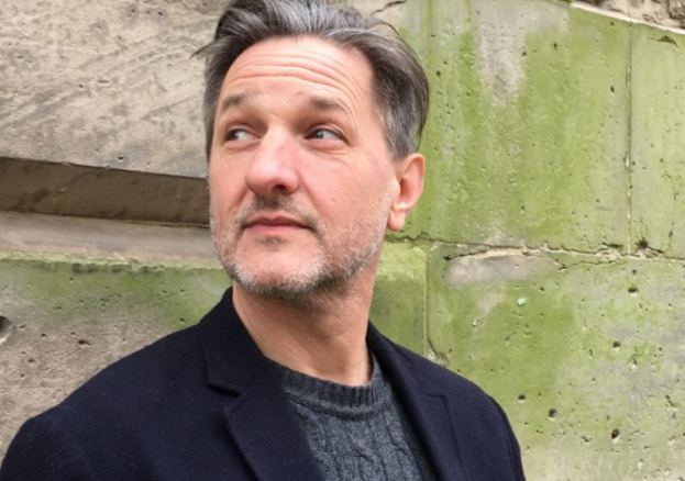 Image of author C. D. Rose looking to the side, wearing a black coat and grey jumper, stood in front of a wall.