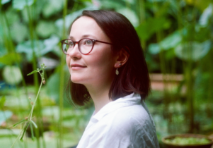An image of author Marianne Brooker stood in front of green fauna, wearing glasses, smiling and looking up away from camera.