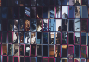 An artwork featuring many rectangular mirrors with coloured frames. In the mirrors are the fractured reflections of a young woman with dark hair, in the act of taking a photograph of two other unidentifiable young women in a street.