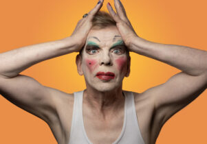 David Hoyle in make-up wearing  whit vest with his hands pulling his face back again an orange backdrop