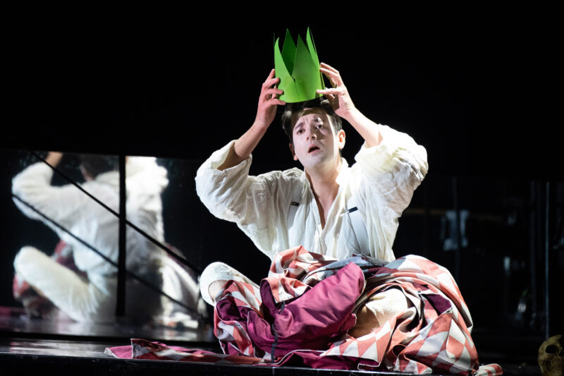 Production image of a man in white pyjamas sat cross-legged on the floor. He is holding a green paper crown to his head. 