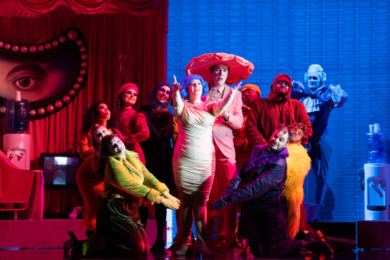 Production image showing cast members in colourful costumes circle round a women in a white dress and blue hair with her arms held out in front of her. 