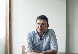A white, middle aged man with short brown hair and wearing a light blue shirt, sits at a round table by a window and against a white wall. He has his arms crossed on the table. 