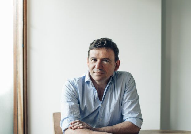 A white, middle aged man with short brown hair and wearing a light blue shirt, sits at a round table by a window and against a white wall. He has his arms crossed on the table. 