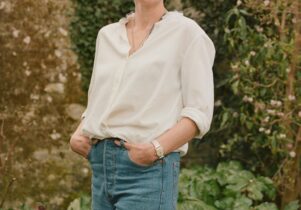 a white middle aged woman stands in front of flower beds and a soft beige stone garden wall with ivy.  she is wearing a white blouse and pale blue jeans and is smiling. She has dark, short hair.