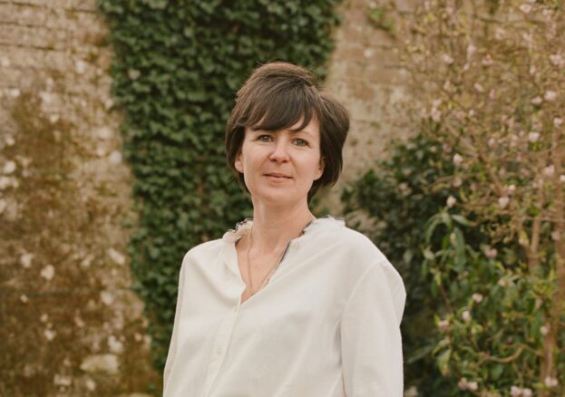 a white middle aged woman stands in front of flower beds and a soft beige stone garden wall with ivy. she is wearing a white blouse and pale blue jeans and is smiling. She has dark, short hair.