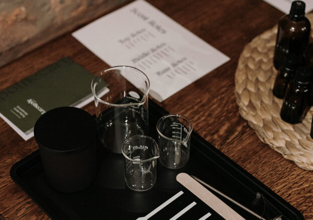 A tray on a wooden table. On the tray there is equipment for candle making such as scent strip, beakers, thermometer, wick and container. 