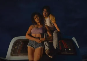 In a dark night, Jackie and Lou face the camera while leaning on a car.