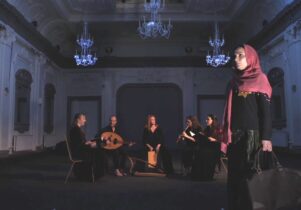 Into the Melting Pot at Manchester Jewish Museum: A photograph showing a theatre stage. On the right side we can see a woman in a pink hijab with a travel bag in her hand. She has a yellow star pinned to her black blouse. She looks concerned. In the background there is a group of 5 musicians playing medieval instruments.