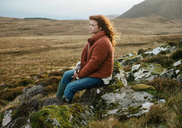 A woman sits on a rock in the countryside. There are green hills and landscape in the back ground.