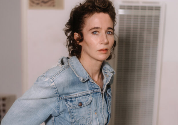 a white woman with swept back culy auburn hairs stares into the camera. she is in a white room near an air conditioning vent and is wearing a faded denim jacket