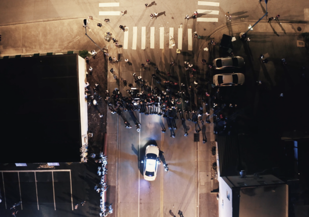 Crowd standing in a street lit by a police car's headlights, captured from above.