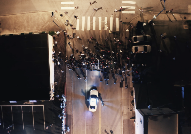 Crowd standing in a street lit by a police car's headlights, captured from above. 