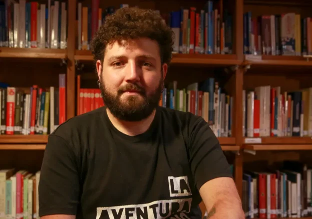 A person - Munir Hachemi - with brown curly hair and a beard sat in front of a book case.
