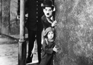 A black and white still from the film THE KID featuring Charlie Chaplin with a child and policeman on a street corner