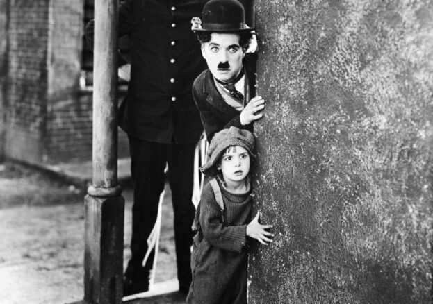 A black and white still from the film THE KID featuring Charlie Chaplin with a child and policeman on a street corner