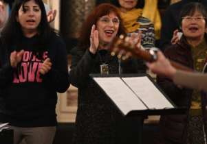 A photograph of people standing in a row and singing together. There are 3 people in the front and 3 people in the background. The people in the front are 3 women. The first person on the left has long dark hair and dark eyes. She is wearing a black hoodie with a print 
