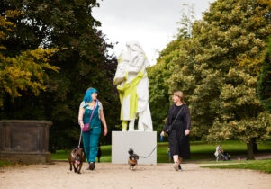 Two people walking their dogs in front of a sculpture