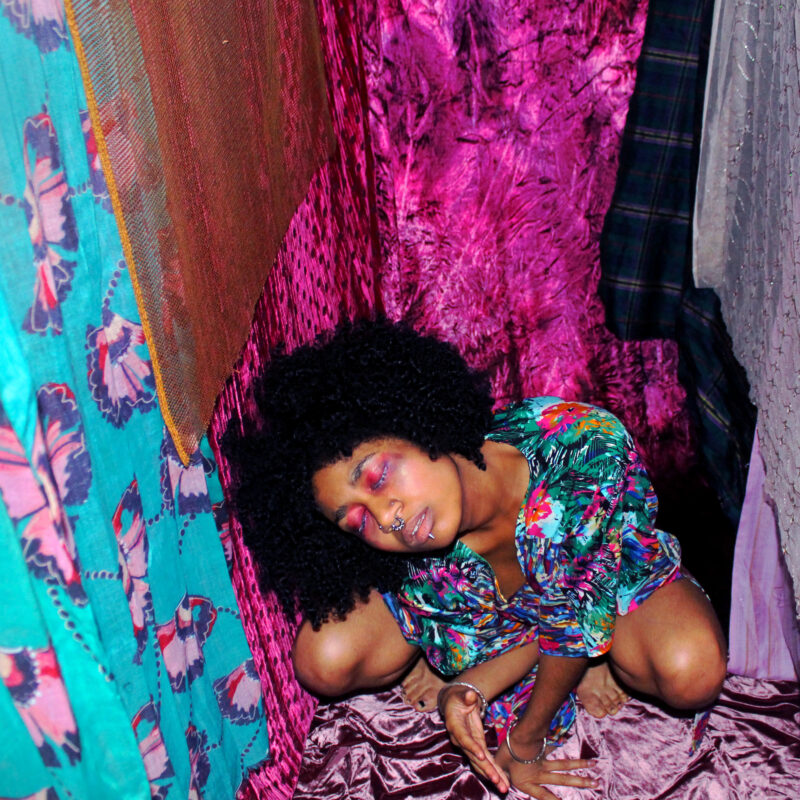 A woman is crouched down around different fabrics with her eyes half closed. She has afro hair and is wearing a bunched up patterned top.