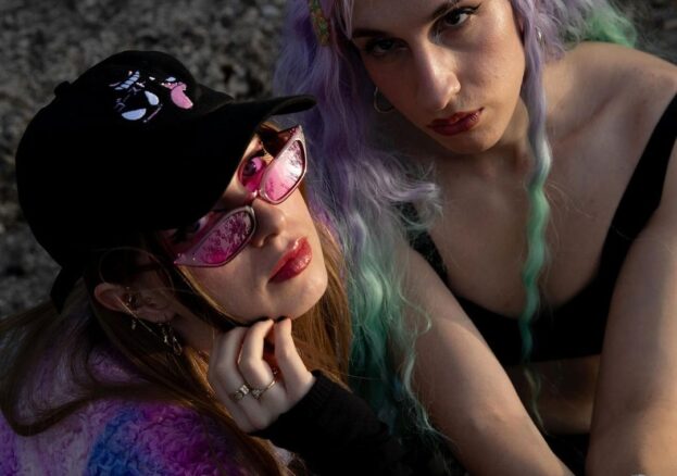 Two women looking up at the camera, one is wearing sunglasses and a cap, holding a phone. The other has long wavy pastel hair.