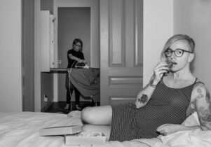Black and white photograph. Woman lays on bed looking at the camera, in the background, a little girl with Downs Syndrome irons.